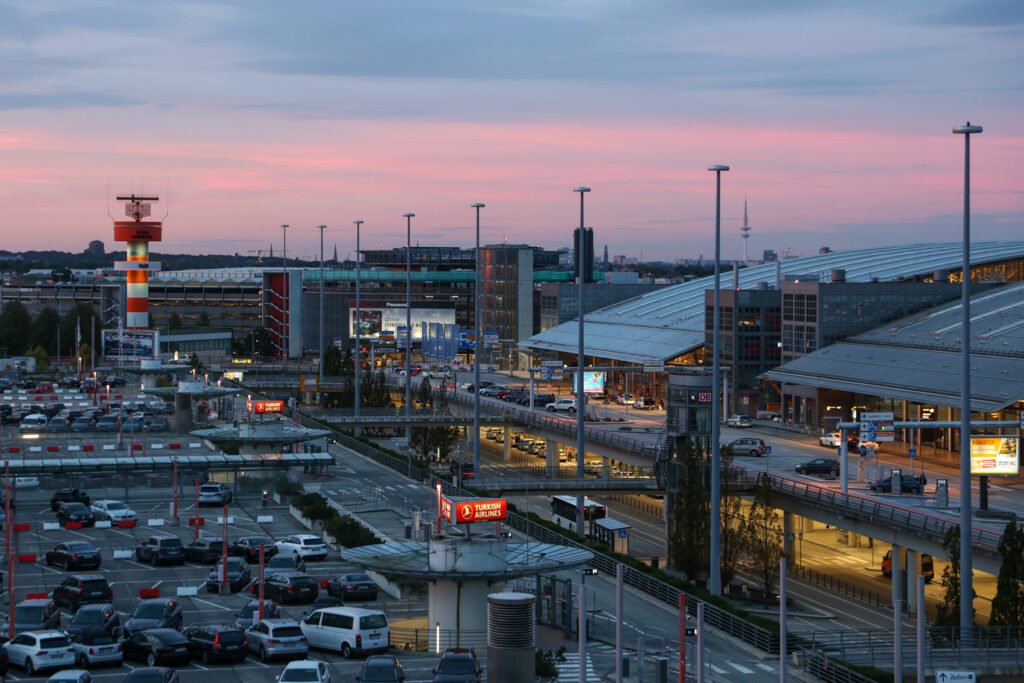 Ein Panorama-Blick auf den Flughafen Hamburg bei Sonnenuntergang, mit vielen geparkten Autos im Vordergrund und dem Terminal im Hintergrund.